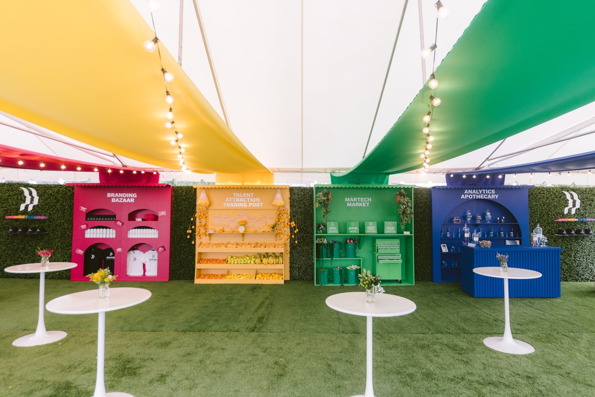 Four brightly colored pop-up booths labeled “Branding Bazaar,” “Talent Attraction Trading Post,” “Martech Market,” and “Analytics Apothecary” arranged under a tent with green turf flooring and white pedestal tables.