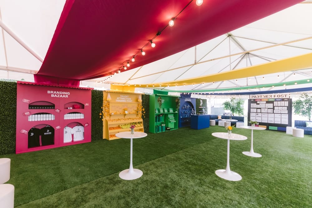 A large event tent with artificial grass, featuring three labeled booths for Branding Bazaar, Talent Attraction Trading Post, and Partner Post, with white cocktail tables arranged in the foreground.