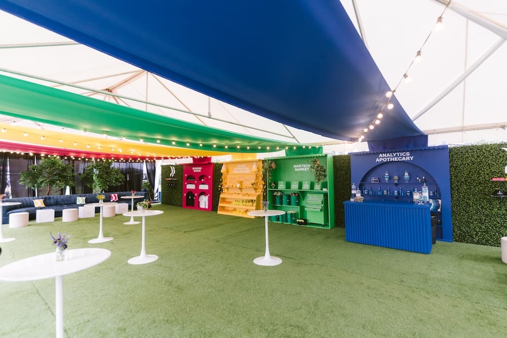 Outdoor event space with a green turf floor, white high-top tables, and colorful booth stations labeled Martech Market and Analytics Apothecary under a rainbow-colored canopy.
