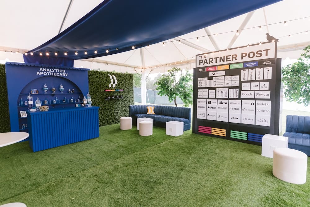 An outdoor lounge area featuring a blue branded “Analytics Apothecary” bar, a dark blue sofa, and a large display board titled “The Partner Post” featuring various company logos.