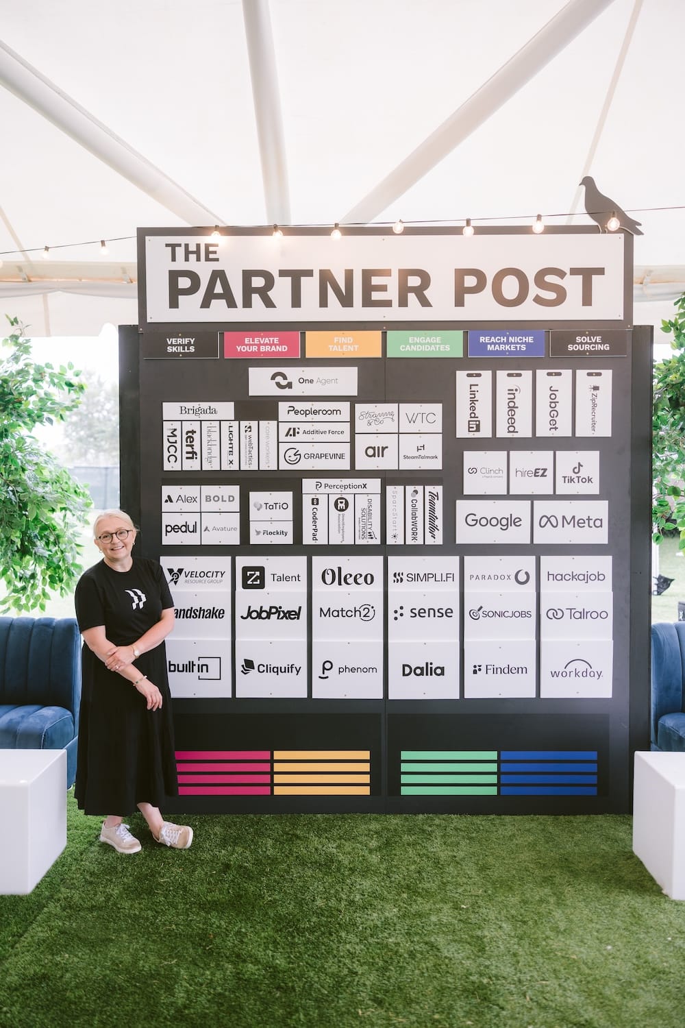 A woman stands next to a large display board titled “The Partner Post,” which features a grid of logos from various companies organized under six categorical headings.