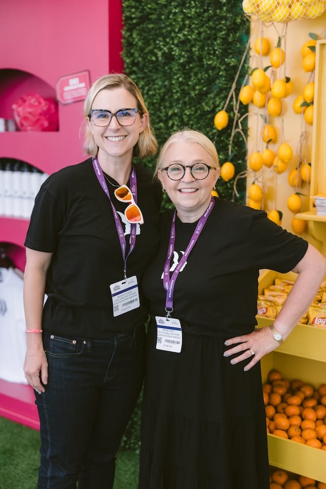 Two women wearing black t-shirts and conference name tags stand in front of a decorative wall display of lemons.