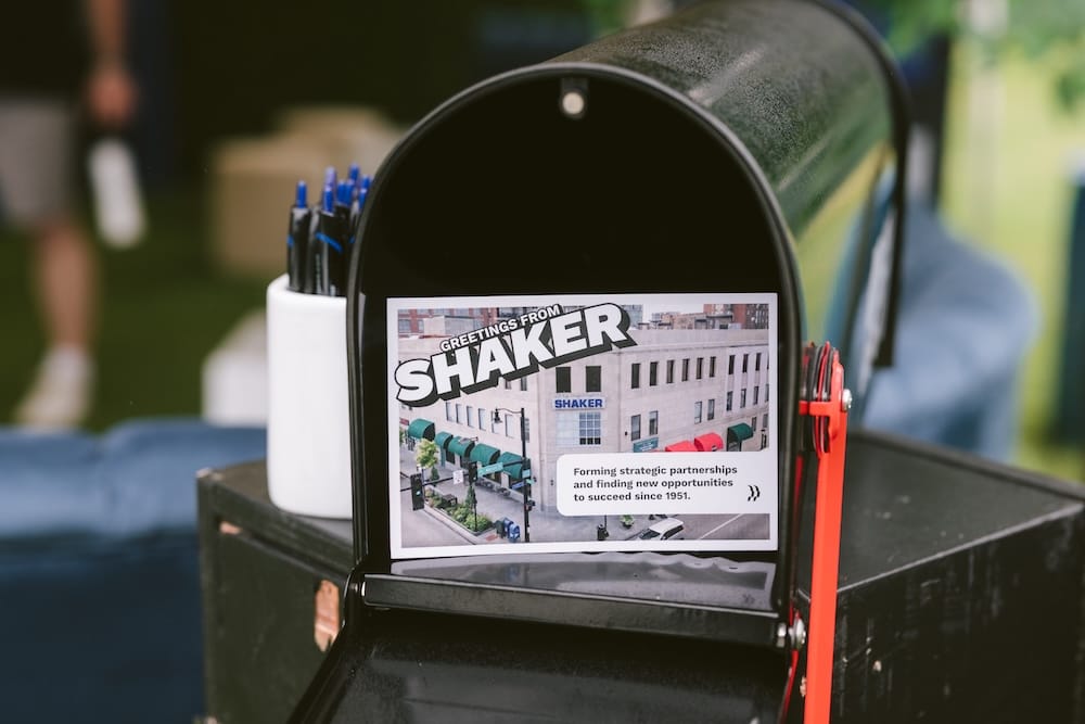 A black mailbox containing a postcard that reads, “Greetings from Shaker: Forming strategic partnerships and finding new opportunities to succeed since 1951.”