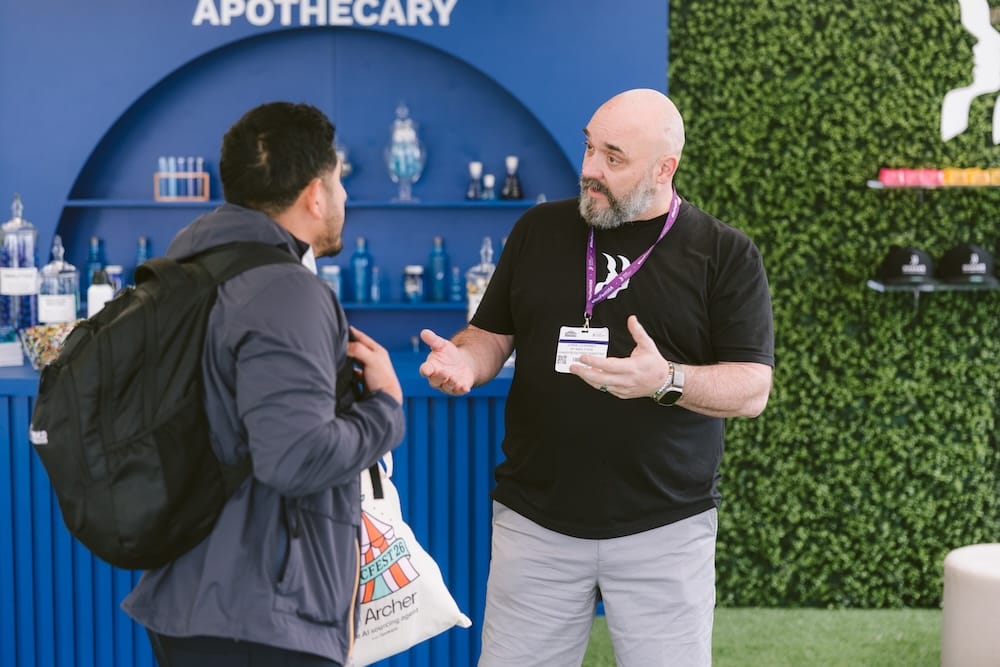 A man in a black t-shirt and lanyard gestures while speaking to another man wearing a backpack in front of an apothecary-themed display.