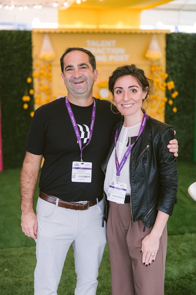A man in a black t-shirt and light-colored trousers stands with his arm around a woman in a white top and black jacket, both wearing conference badges at an event.
