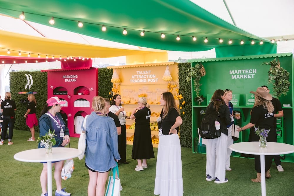 Attendees walk between three branded booths labeled Branding Bazaar, Talent Attraction Trading Post, and Martech Market at an outdoor event under string lights.