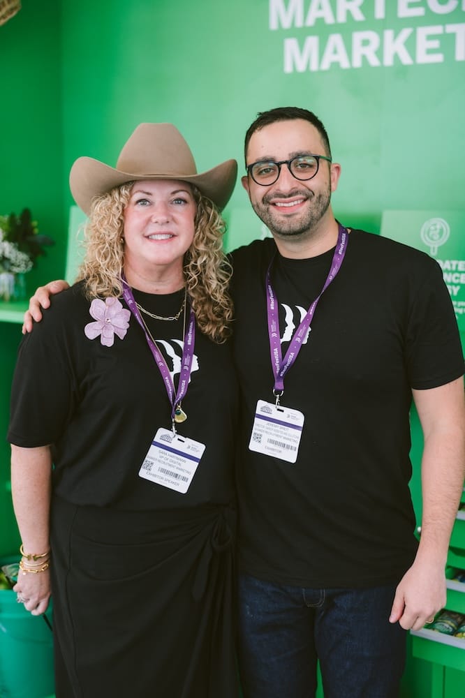 A woman wearing a cowboy hat and a man wearing glasses stand side-by-side in front of a green backdrop, both wearing black shirts and event name tags.