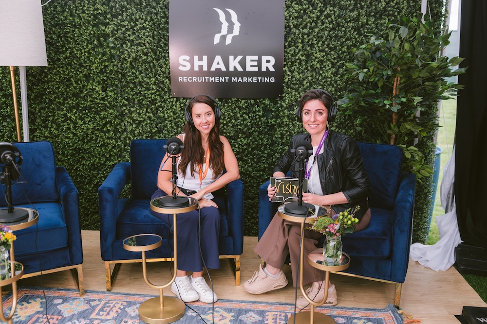 Two women wearing headsets sit in blue armchairs in front of a Shaker Recruitment Marketing sign, preparing to record a podcast.