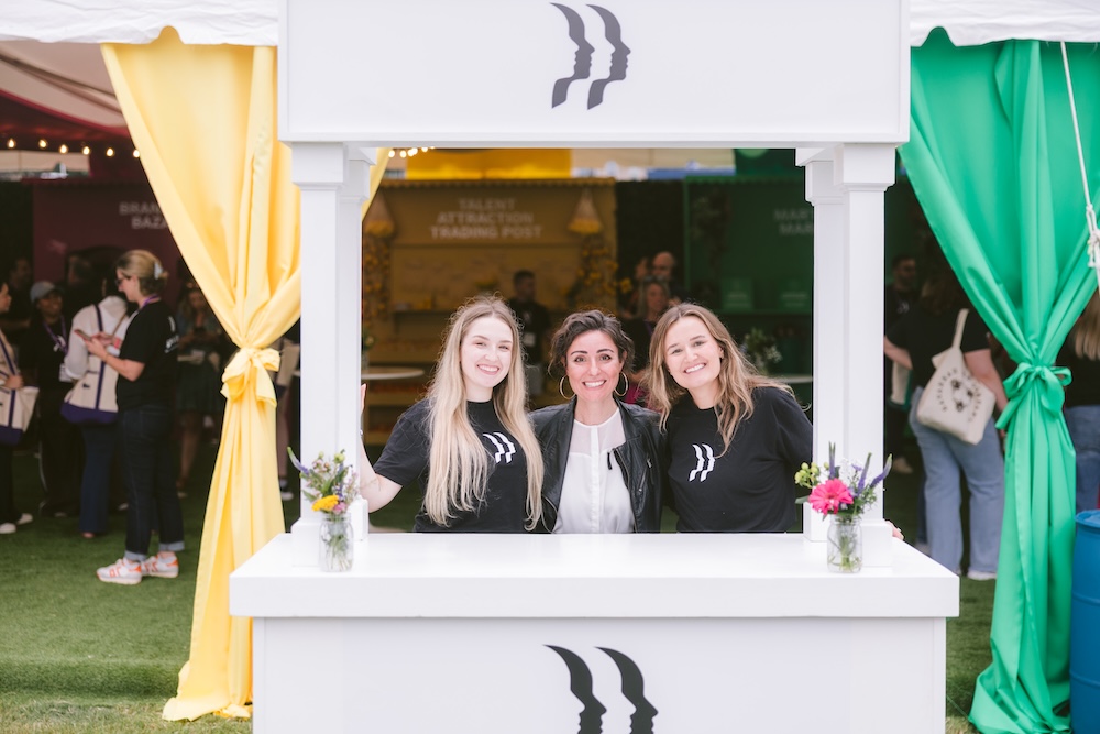 Three women standing behind a white event booth, each wearing a black t-shirt with a double-silhouette logo.