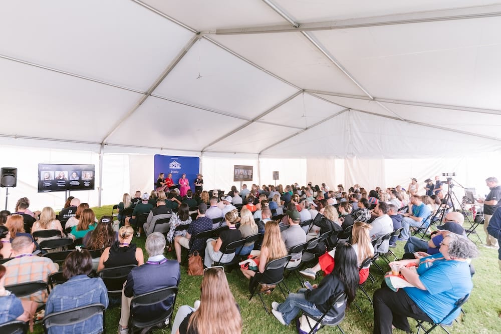 A large audience sits in folding chairs under a tent, watching two speakers on a stage with a branded backdrop.