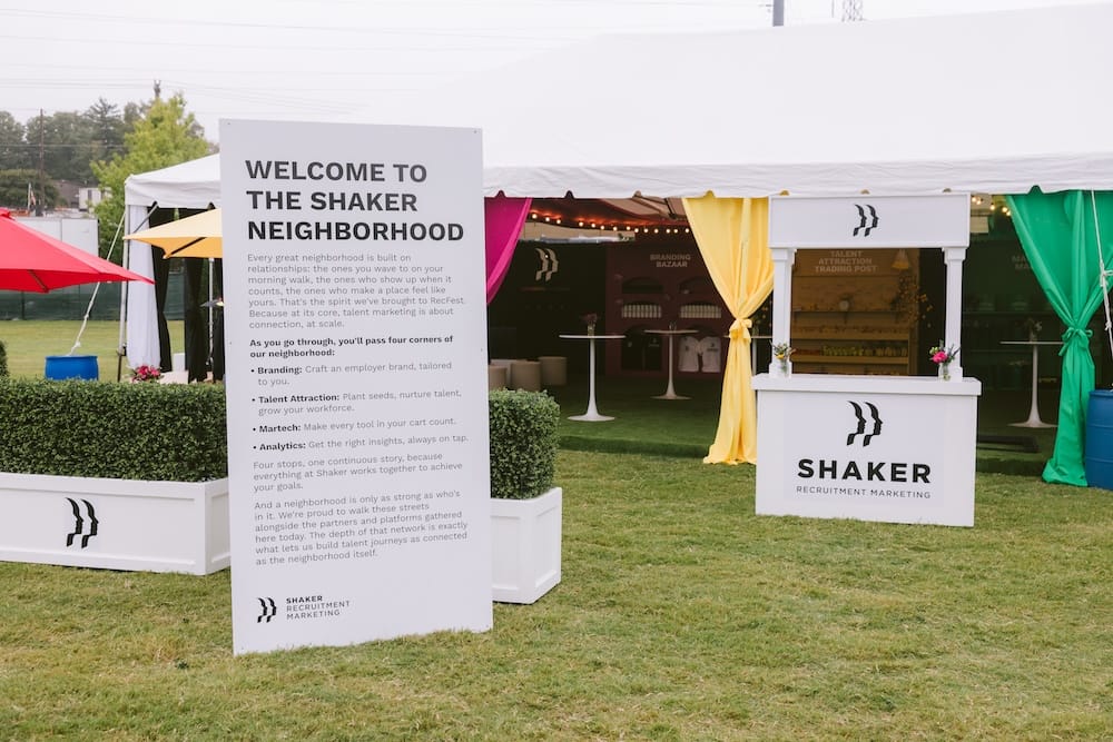 A Shaker Recruitment Marketing event display with a large sign detailing the “four corners of our neighborhood” next to a white information booth with colorful fabric drapes.