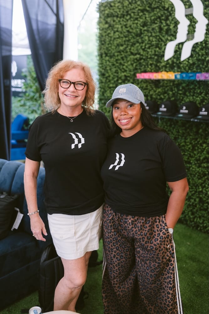 Two women smiling side-by-side, both wearing black t-shirts featuring a white double-profile logo.