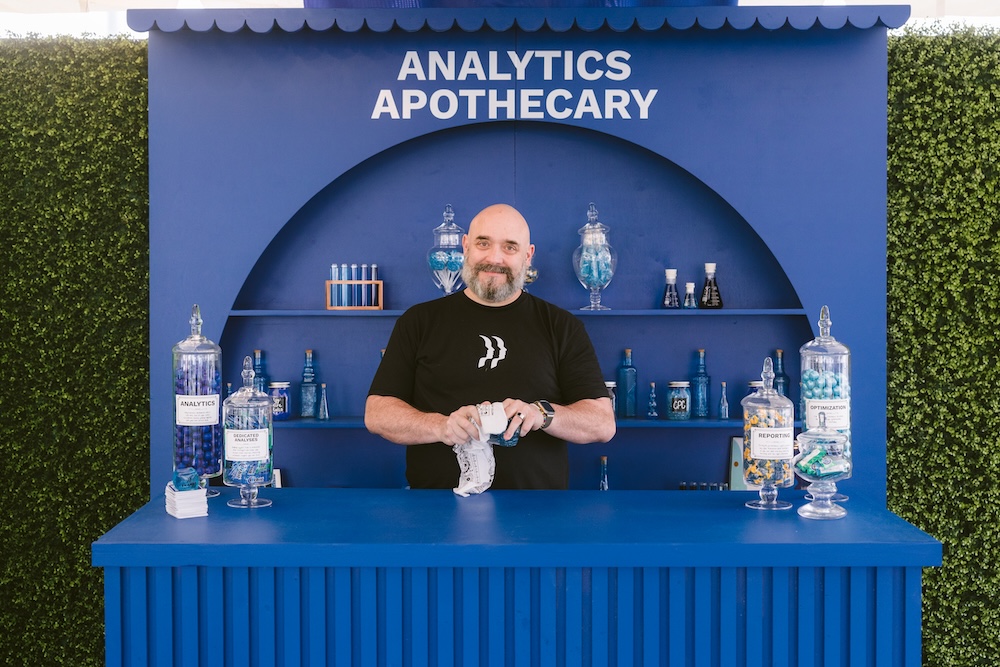 A man with a beard stands behind a blue “Analytics Apothecary” booth filled with glass jars containing blue candies.