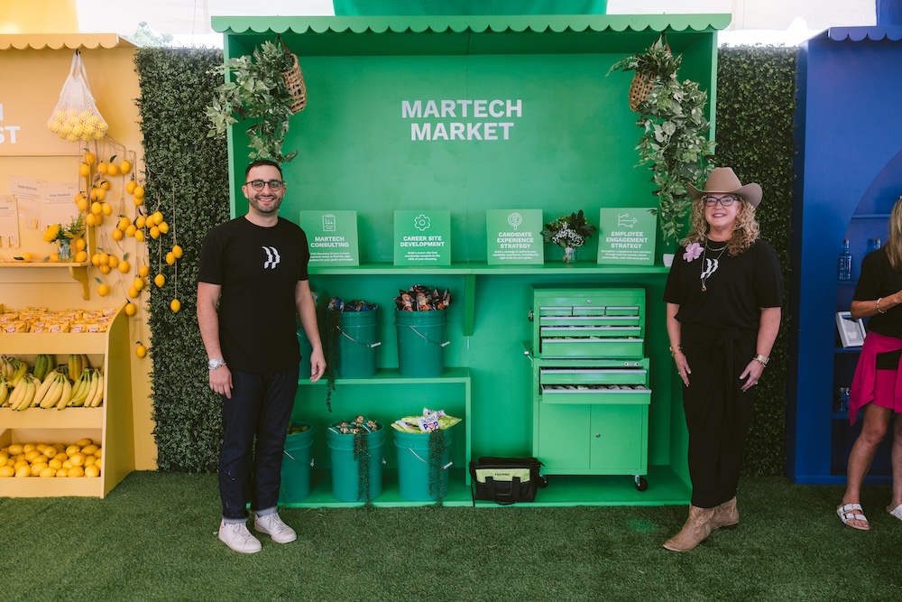 A man and a woman in black branded shirts stand in front of a green Martech Market display booth with four informational signs, snack-filled buckets, and a green metal tool chest.
