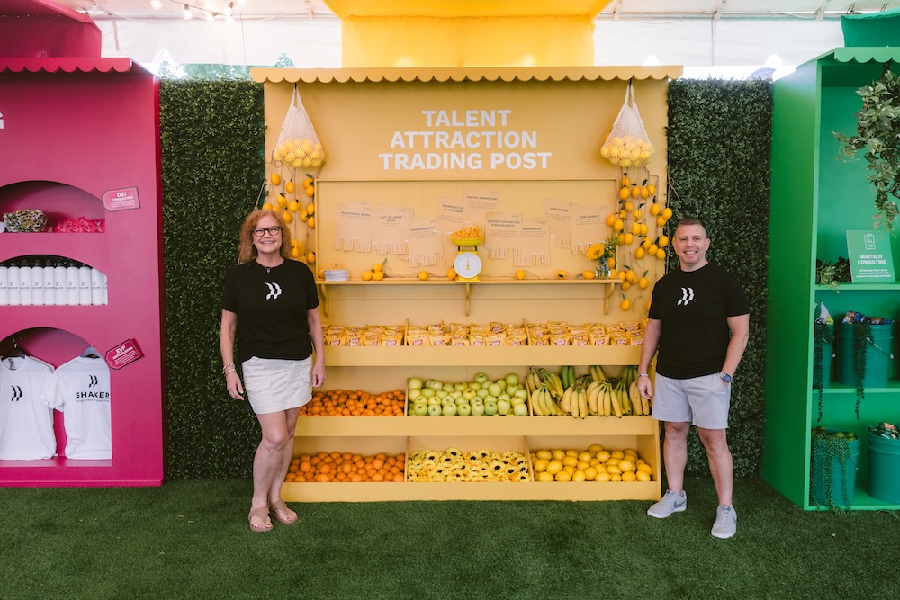 A man and a woman in branded black t-shirts stand in front of a yellow “Talent Attraction Trading Post” booth filled with fruit and marketing display materials.