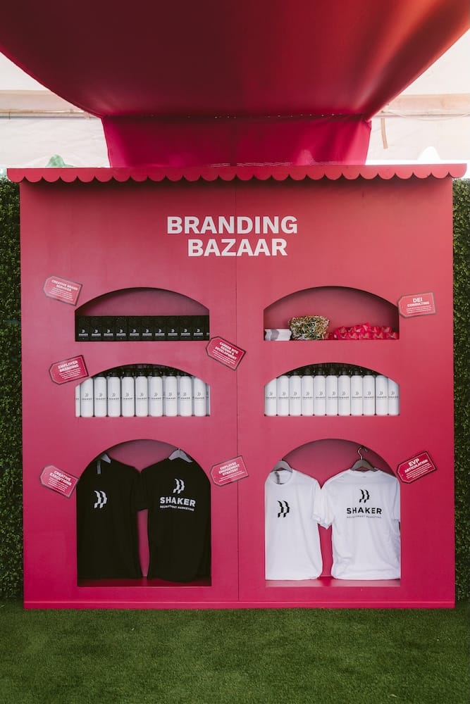 A pink, open-front display booth labeled “Branding Bazaar” featuring shelves of white water bottles and clothing with the Shaker Recruitment Marketing logo.