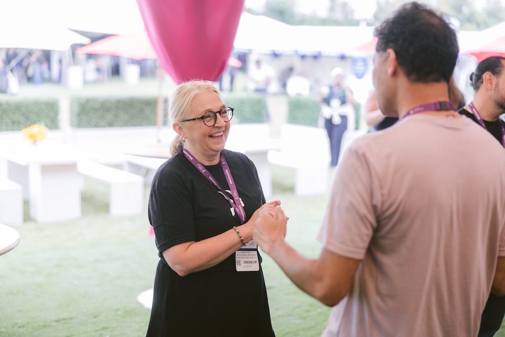 A woman in a black shirt and conference lanyard laughs while talking to a man at an outdoor event.